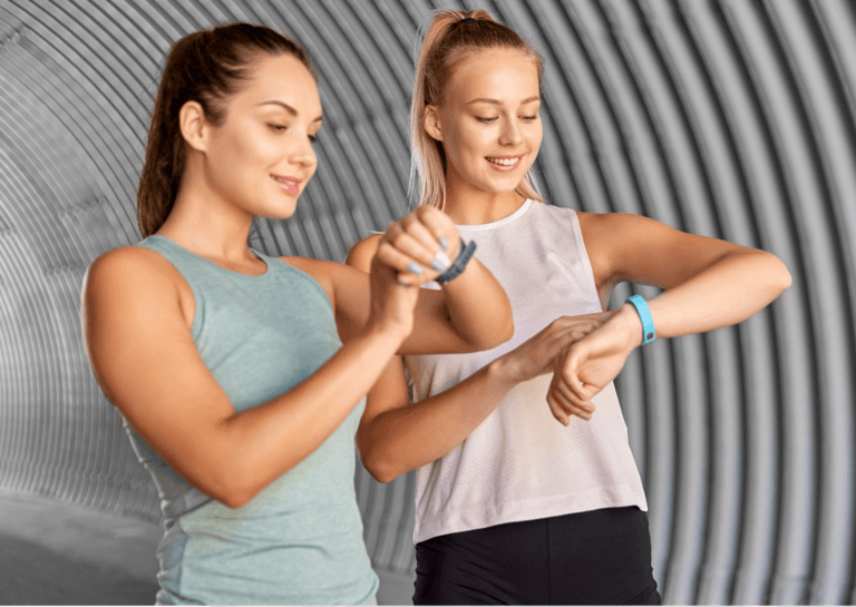 Two young women with ponytails, one brunette in a light blue-green sleeveless top and one blonde in a pink sleeveless top, happily checking and using their fitness trackers in a tunnel with a textured, ribbed interior.