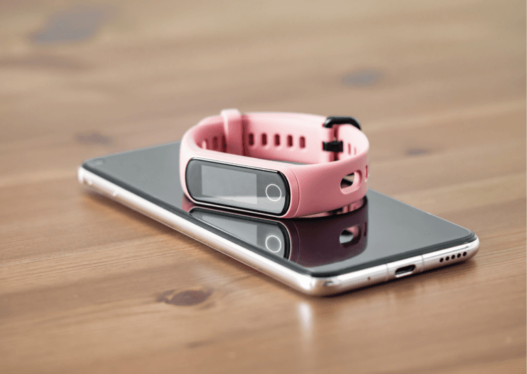 Pink fitness tracker watch resting on a smartphone, both placed on a wooden surface, highlighting their sleek designs and tech features.