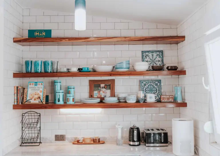 Three wood-colored floating shelves evenly spaced above a white wall, displaying various decorative items and placed above a countertop.