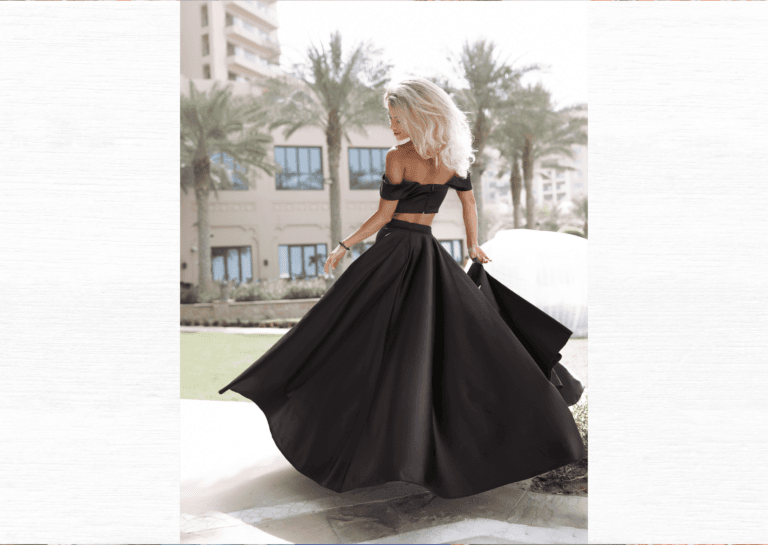 Young woman in a formal dress with a wide bottom, photographed from behind as she looks back at her gown, standing amidst tall palm trees and a building in the background, showcasing her full blonde hair.