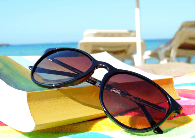 Close-up of black sunglasses resting on a colorful beach read under a sun umbrella, with two lounge chairs blurred in the background and the sea visible.