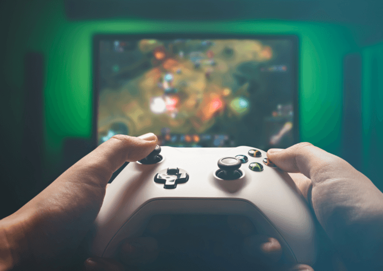 Close-up of a person holding a white Xbox gaming console, with a TV displaying the game in the background and a softly illuminated light green backdrop.