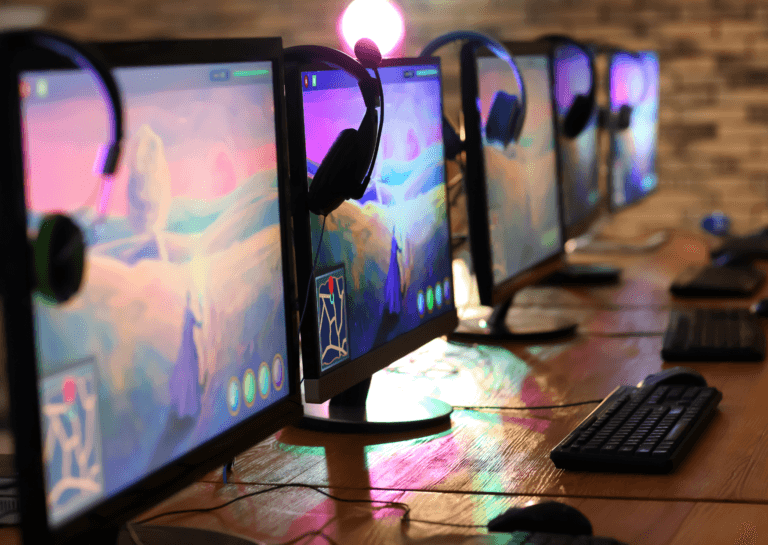 Five gaming PCs arranged diagonally on a long brown table, featuring vibrant backgrounds and headsets draped over them in a gamer setup.