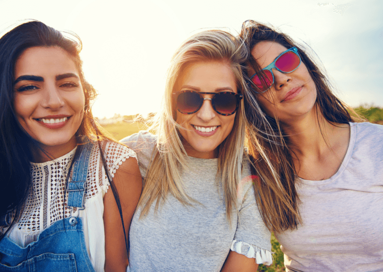 A group of three young women with long hair, smiling in the sunlight. The woman on the left has black hair and wears a denim overalls, the blonde woman in the middle wears brown sunglasses, and the brunette on the right leans her head on the blonde woman, wearing red-lens sunglasses with a light blue frame. Only the upper half of their bodies is visible.