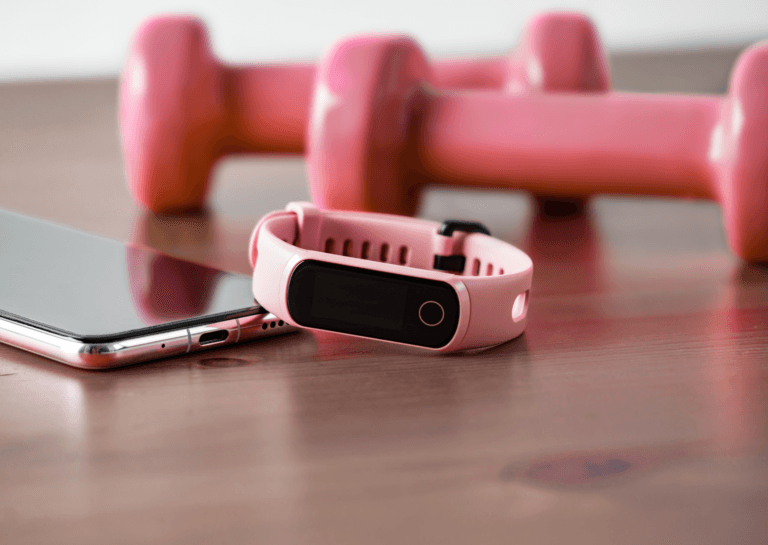 Pink smartphone with a Fitbit smartwatch displayed alongside light dumbbells, all set on a wooden surface, creating a stylish fitness-themed arrangement.