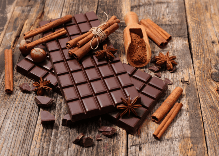 Gourmet delicatessen featuring chocolate bars cut into squares, bundled cinnamon sticks, star anise, and a cinnamon scoop filled with cinnamon, elegantly arranged on a wooden surface.