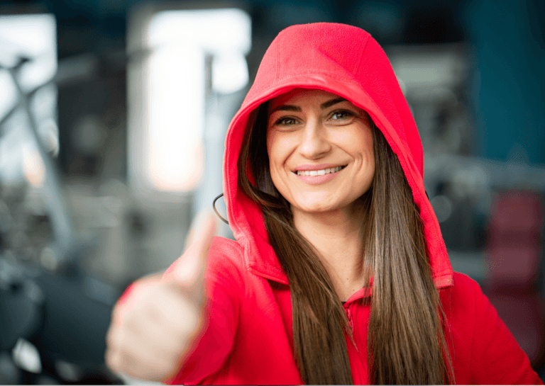 Brunette woman wearing a red hoodie with the hood up, smiling and giving a thumbs up; her long brown hair peeks out from the hoodie against a blurred gym-like background.