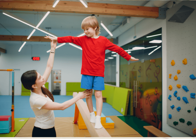 Young boy with medium-length blond hair, wearing a red long-sleeve shirt and blue shorts, balancing on a gymnastics beam in a gym. He looks toward a young female instructor with long brown hair in a ponytail, wearing a light shirt. Background shows gym mats and a climbing wall.