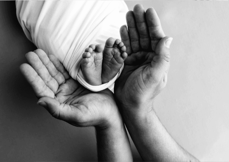 A close-up image capturing only the feet of a swaddled baby, resting above two open adult hands positioned underneath. The contrasting tones emphasize the tenderness and connection in this intimate moment.