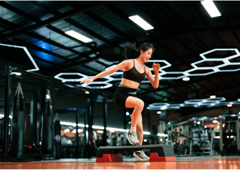 Young woman in a stylish gym with her hair in a ponytail, wearing a black crop top and shorts, jumping over a bench from left to right; mirrored walls and various fitness equipment are visible, filmed from the floor with ceiling lights in view.