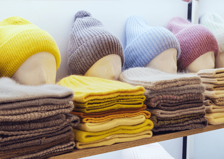 Five winter hats in yellow, brown, blue, pink, and white displayed on a mannequin head, with a stacked assortment of the same colored hats arranged in front. The scene is set on a wooden plank against a white background, creating a colorful and cozy winter vibe.