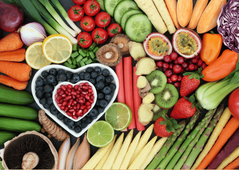 An assortment of colorful fruits and vegetables including bananas, tomatoes, lemons, cucumbers, kiwis, onions, green asparagus, strawberries, radishes, and bell peppers arranged together, with a heart-shaped white porcelain bowl filled with blueberries in the center-left and a smaller heart-shaped bowl containing red fruits nested inside.