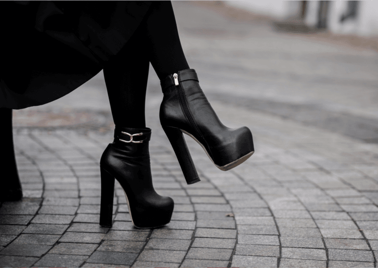 Black high-heeled shoes with gold detailing on the side, worn by a woman sitting outdoors on the street with her legs crossed, showcasing the square cobblestones beneath.