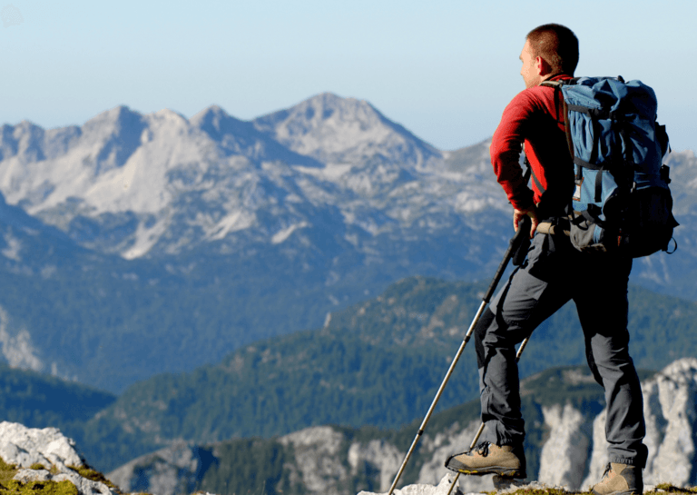 Man in a red sweater and large blue backpack, wearing hiking pants, gazing at the mountains while holding trekking poles and wearing hiking shoes.