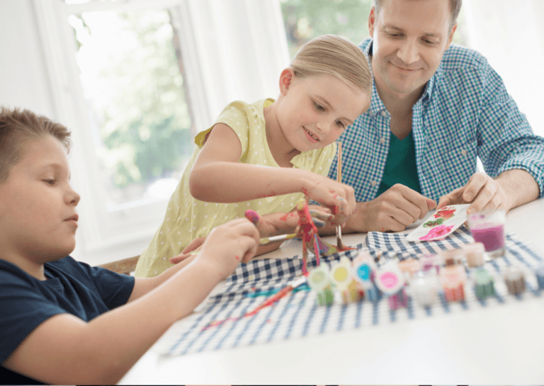 A boy and girl around 10 years old sitting at a white table with their father, surrounded by paintbrushes and paint on a checkered white and blue cloth. They are focused on their artistic activities, enjoying a fun and creative time together.