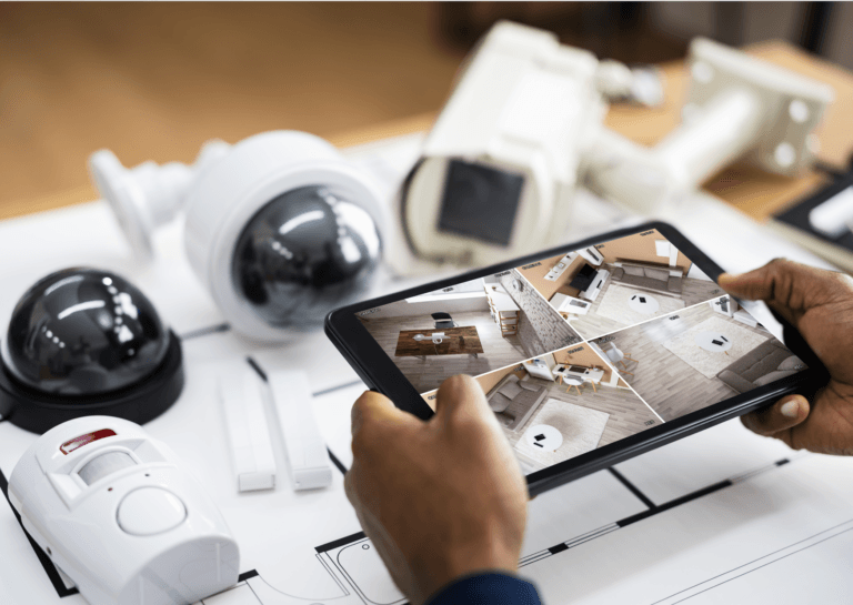 A man’s hands holding a tablet displaying four security camera feeds, with several unconnected white security cameras on the table behind it.