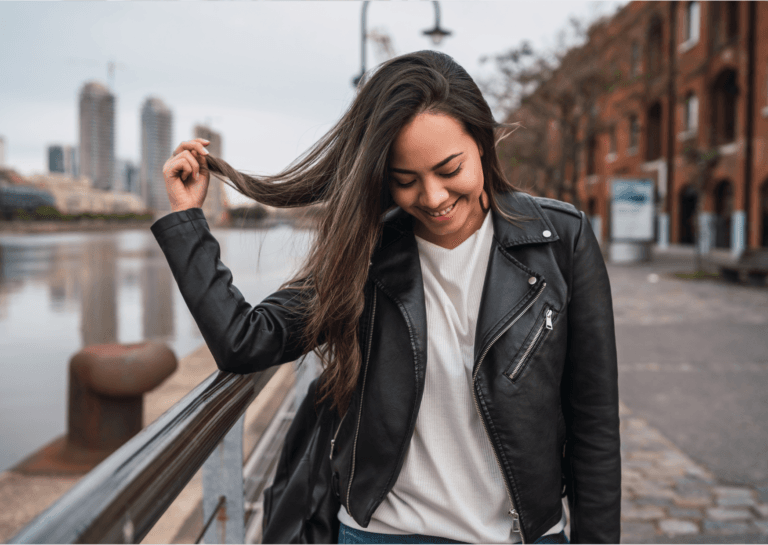 Woman with long dark hair, smiling and looking down at the street, wearing a black leather jacket and holding a strand of hair with one hand, standing between the canal and the street.