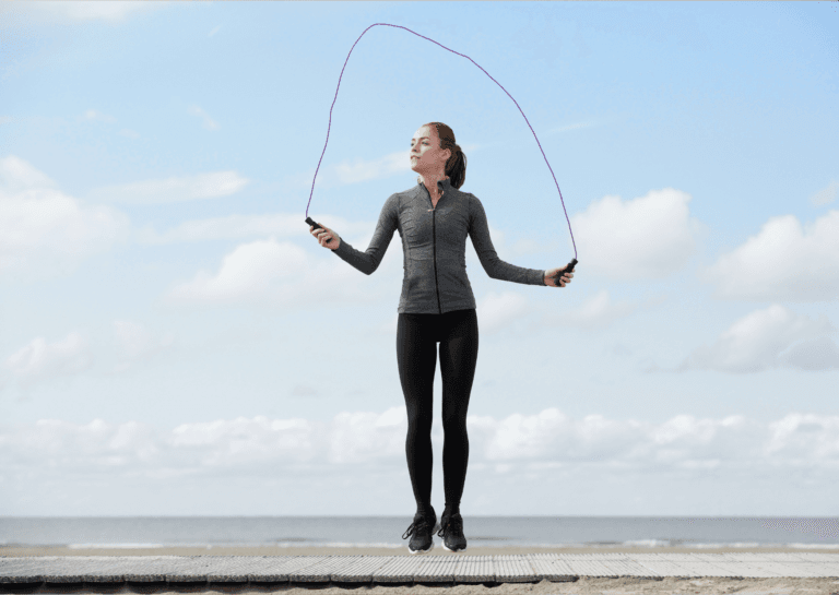 Woman jumping rope on the beach, with the rope above her head, captured mid-air while looking to the left; the sea is visible in the background along with a boardwalk running from left to right, and she is dressed in a long-sleeved athletic top, leggings, and sports shoes.