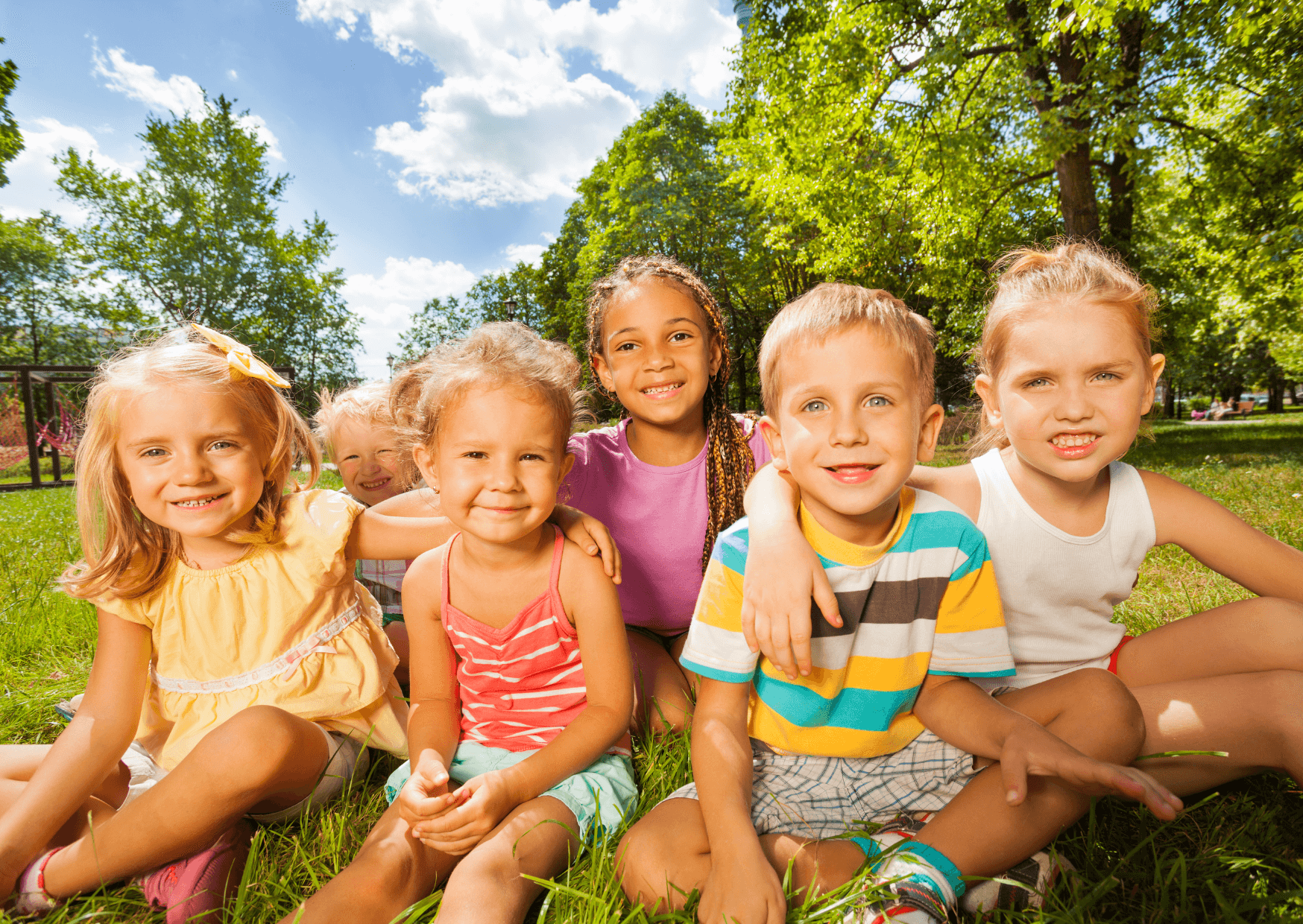 Six children aged 4 to 7 sitting on a sunny grass field, smiling happily for the photo.