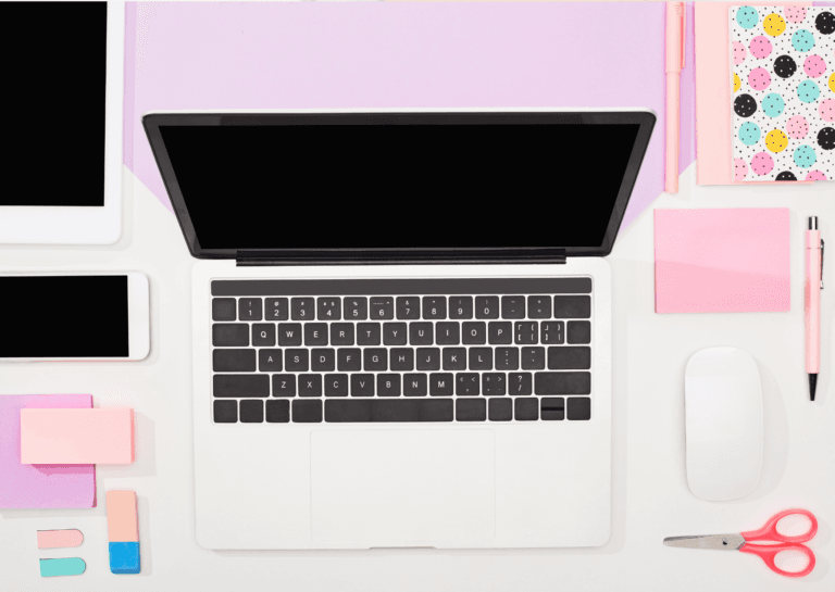 Top view of an open laptop on a white desk, with a white phone, tablet, pink sticky notes, eraser, pink notebook, scissors, and pink marker arranged neatly around it.