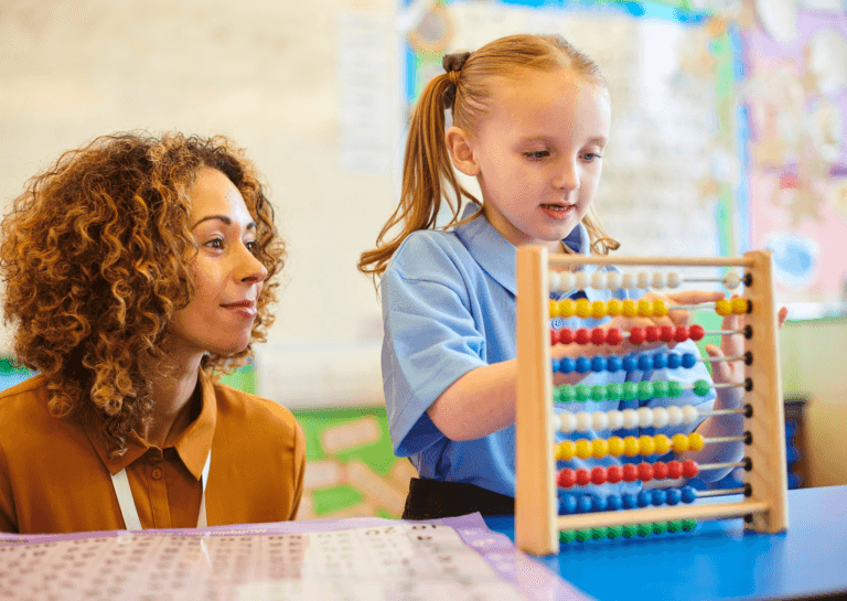 Young girl around 5 years old playing with an abacus, while an adult woman with brown shoulder-length curly hair crouches beside her, laughing and observing like a teacher in a classroom setting.