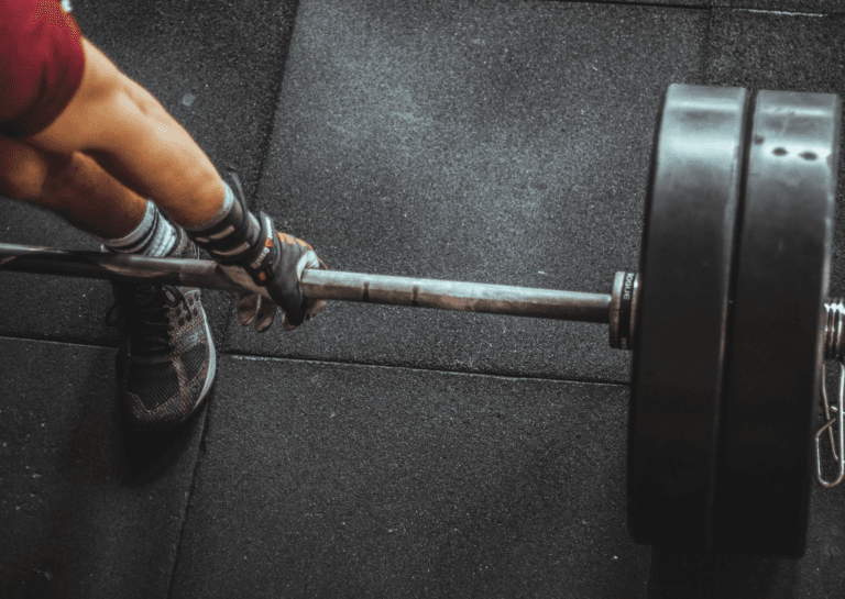 Person wearing lifting gloves positioned to lift a heavy barbell with both hands, though only one hand and foot are visible on square fitness mats.