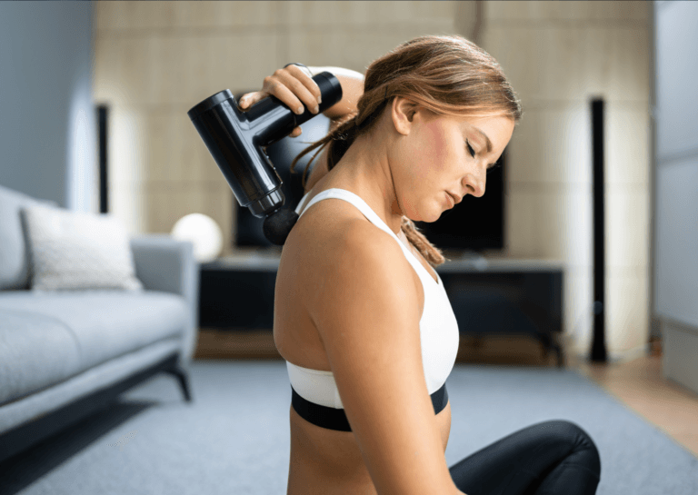 A woman sits cross-legged on the floor in a white sports top, holding a massage gun behind her and pointing it at her back, eyes closed, in front of a couch.