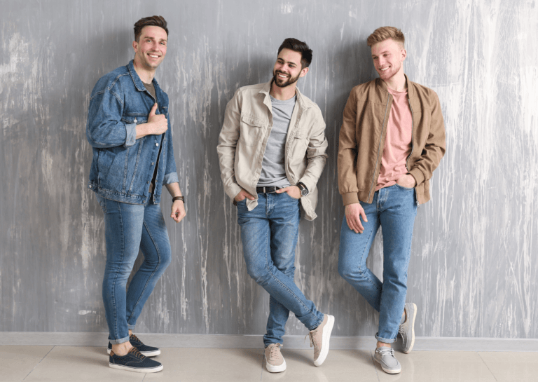 Three well-dressed men in casual outfits, each wearing jeans and a small blazer. Two are looking down while one smiles at the camera, all laughing against a light gray background.