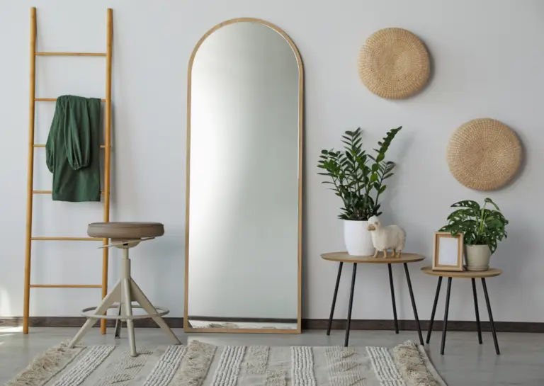 Round-topped standing mirror leaning against a white wall, accompanied by a decorative ladder and stools with plants, adding a fresh touch to the living space.