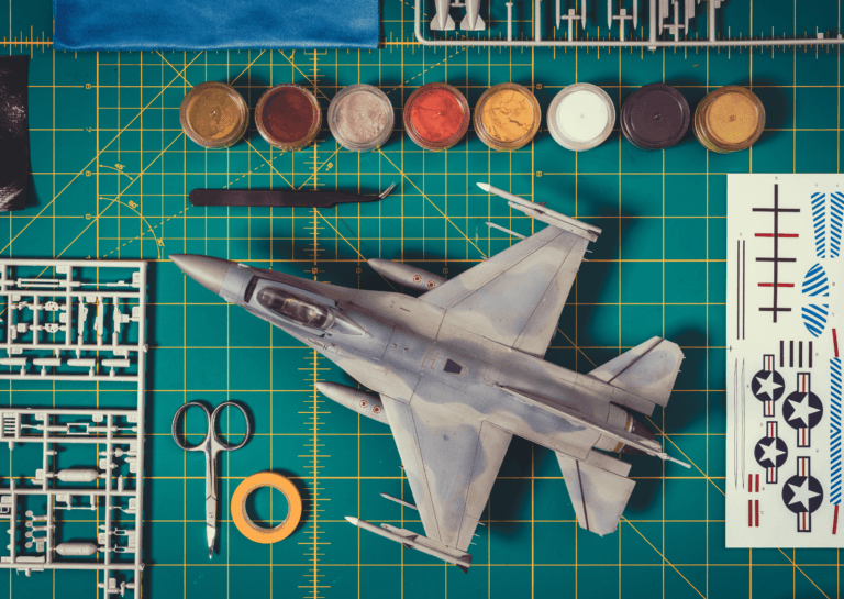 Model kit of a jet fighter viewed from above, showcasing the entire aircraft alongside various parts and tools from the kit, all laid out on a green gridded board that accompanies the kit.