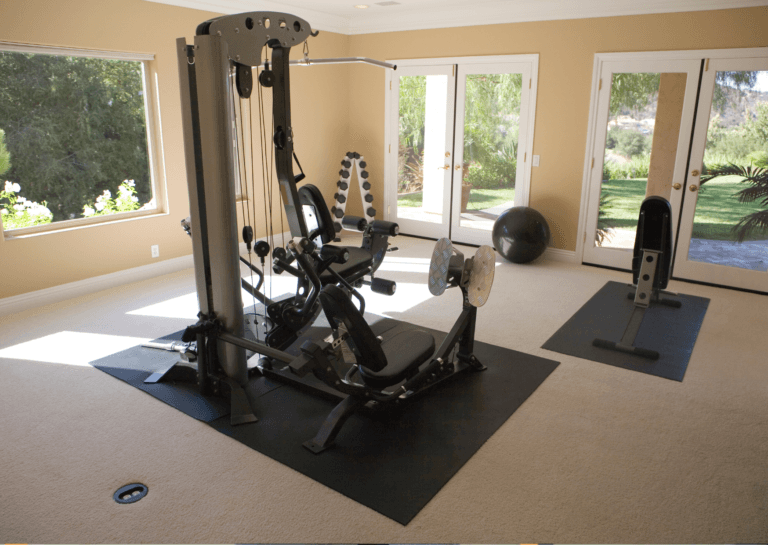 Multistation home gym positioned in the center of a room on a black fitness mat, with dumbbells in the corner on a rack, a fitness ball, and a separate workout bench on the mat. Sunlight streams in during the day, revealing a beautiful green garden visible through the windows.