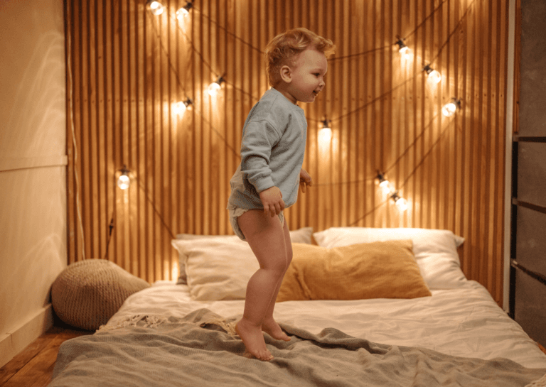 Toddler standing on a single bed, wearing a diaper and a blue shirt, with a vertically striped wooden wall and decorative lights behind the pillows.