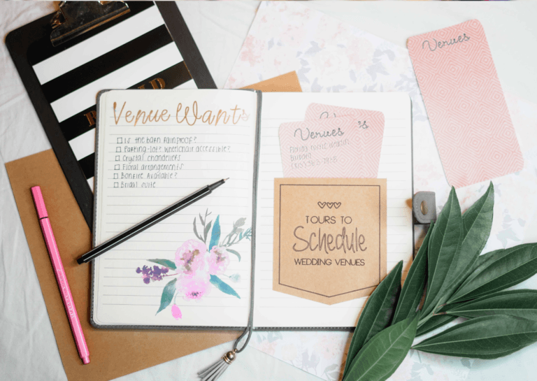 Overhead view of school organizers for planning, featuring an open notebook with the text 'tours to schedule wedding venues' visible inside. Beside it is a notepad holder and a sprig with green leaves, along with a closed pink marker, all arranged on a small desk. This layout showcases a creative and organized workspace.