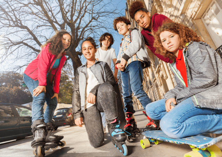 Four girls and two boys crouching or bending down, all on skates, with one girl holding a skateboard. They are smiling at the camera positioned on the ground, capturing a fun moment of their outdoor adventure.