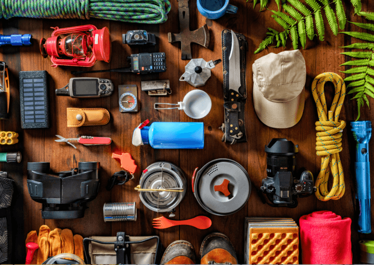 Outdoor adventure gear spread across a brown table, featuring knives, a cap, rope, gloves, a fern, a camera, a percolator, a lamp, an axe, a walkie-talkie, binoculars, solar panel, and shoes.