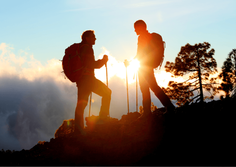 Two hikers standing on a sloped mountain during sunset, with the setting sun peeking through the clouds behind them. The golden light creates a beautiful silhouette of the hikers, surrounded by a stunning backdrop of clouds and a serene evening sky.