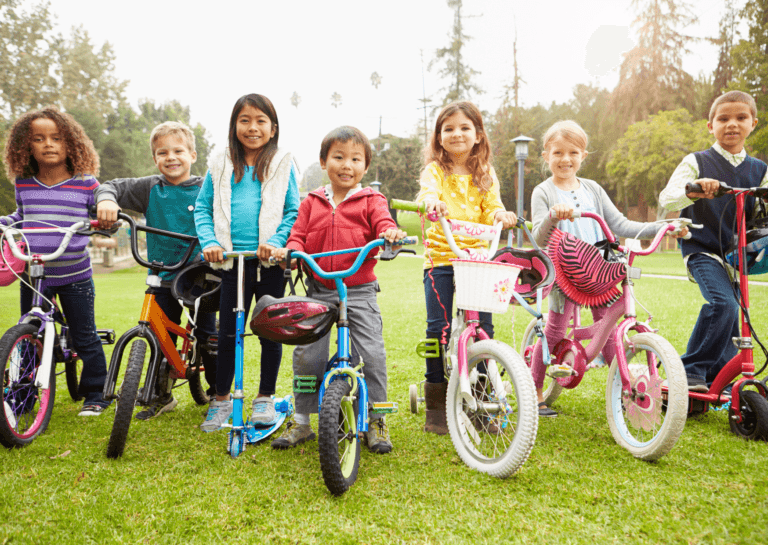 A group of seven smiling kids sitting on bicycles, with one child on a scooter, all on the grass. They are joyfully looking at the camera, capturing a fun outdoor moment.