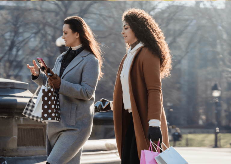 Two young adult women with long hair walking outside in long gray and brown winter coats, each holding two shopping bags in their hands, enjoying a day out.