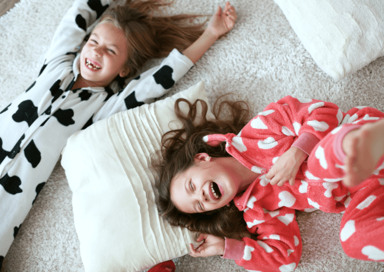 Two young girls around 10 years old, laughing joyfully while lying on a light-colored, plush rug. One girl wears a black and white cow-patterned onesie, and the other is in a red onesie with white hearts, both sporting long dark brown hair.