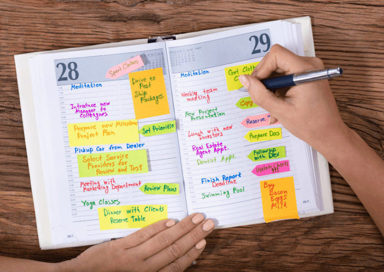 Open planner displaying the 28th and 29th of the month, filled with colorful notes and sticky notes, as a woman with polished nails writes in it with a pen, seen from her perspective with only her arms visible.