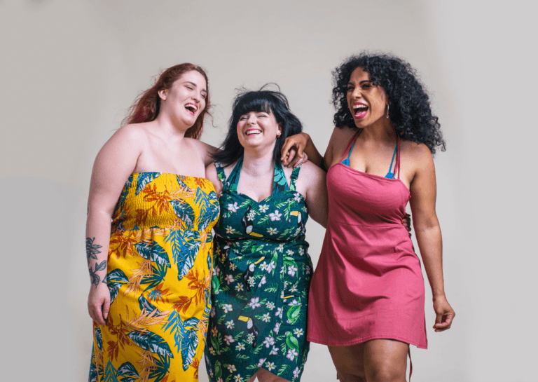 Three plus-size young women holding each other, smiling with their eyes closed in a joyful pose, wearing summer dresses in orange, green, and reddish hues.