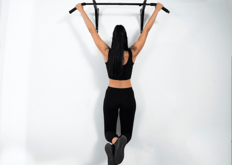 Woman with long dark hair in a ponytail hangs from a pull-up bar, wearing a black top and leggings, legs extended with ankles crossed against a white wall background.