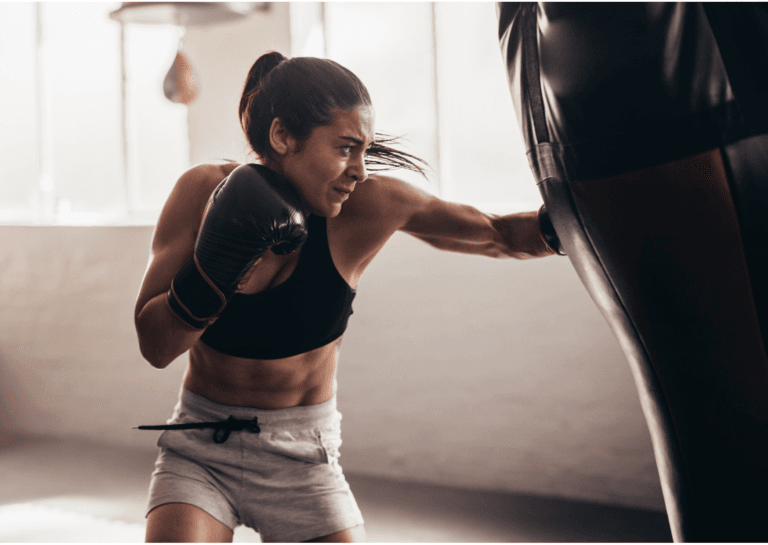 Boxing woman in a black top and short gray shorts, striking a large punching bag that is visibly swinging, positioned in a brightly lit room with sunlight streaming through the window; the woman stands in the center, with part of the moving bag visible on the right.