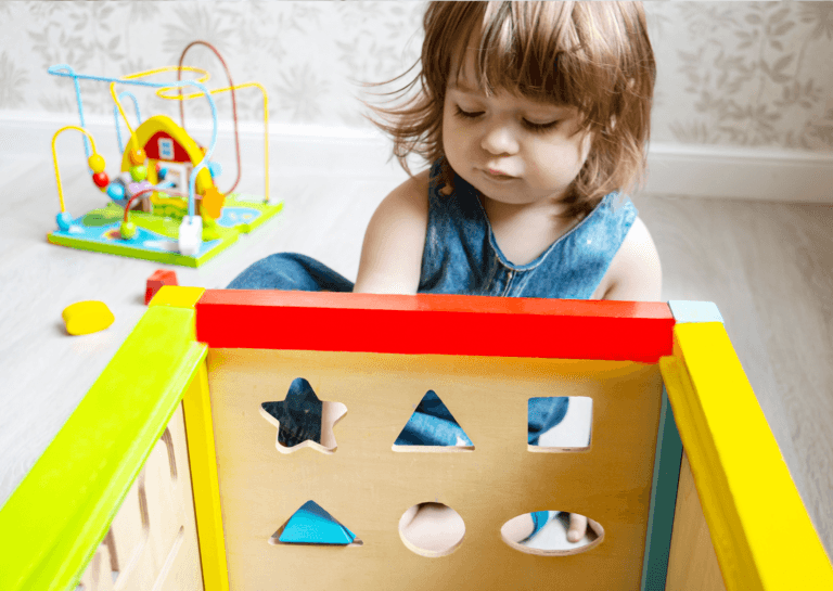 Toddler girl playing with a puzzle shape sorter in a colorful bedroom filled with various toys in the background.