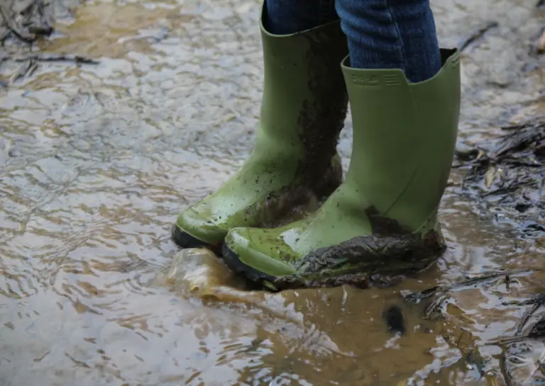 Green rain boots for men, shown in a close-up as someone stands in a puddle of muddy water.