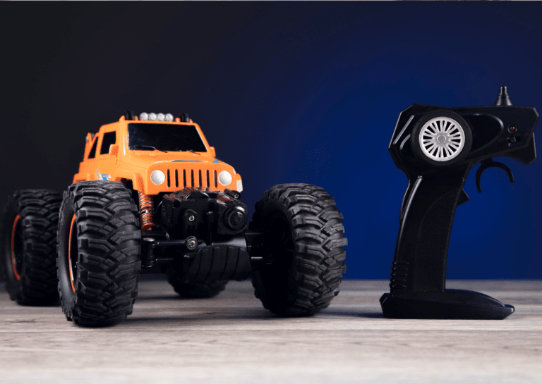 Orange remote-controlled monster truck with large wheels on a light brown table, positioned next to its controller.