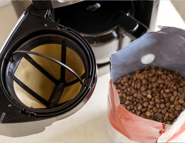 Top-down view of a coffee maker with an empty reusable coffee filter pod inserted, alongside an open bag of coffee beans ready for use.