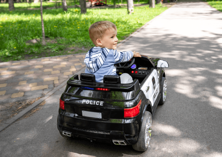 Toddler riding in a police ride-on car on the street, looking to the right with grass and trees visible at the roadside.