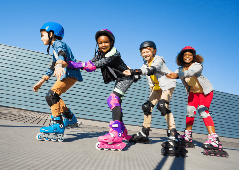 Four young children skating in a line, holding each other by the hips while moving from left to right. They wear colorful clothing and complete protective gear, including helmets.