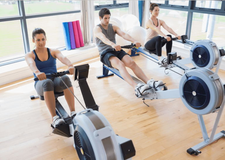 Two young adult women and one young adult man in the center, exercising on rowing machines in the corner of a bright room surrounded by windows, all dressed in athletic clothing.